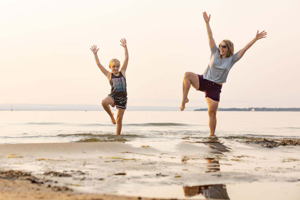 Mentee and Mentor reach for the sky and balance on one leg at the beach, with their ankles in the water.