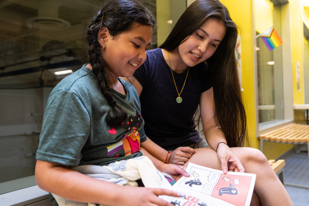 A female mentor with long dark hair reads to a young female mentee with long dark hair with a book on her lap.