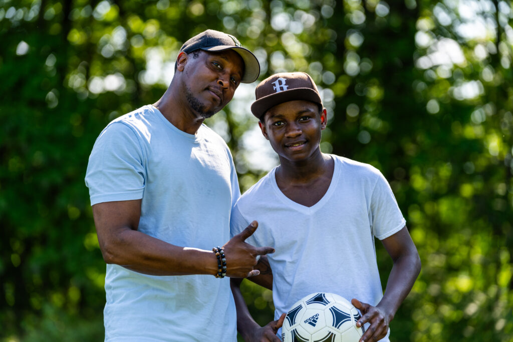 A male adult mentor and his mentee stand side by side in an outdoor setting. Mentee holds a soccer ball.